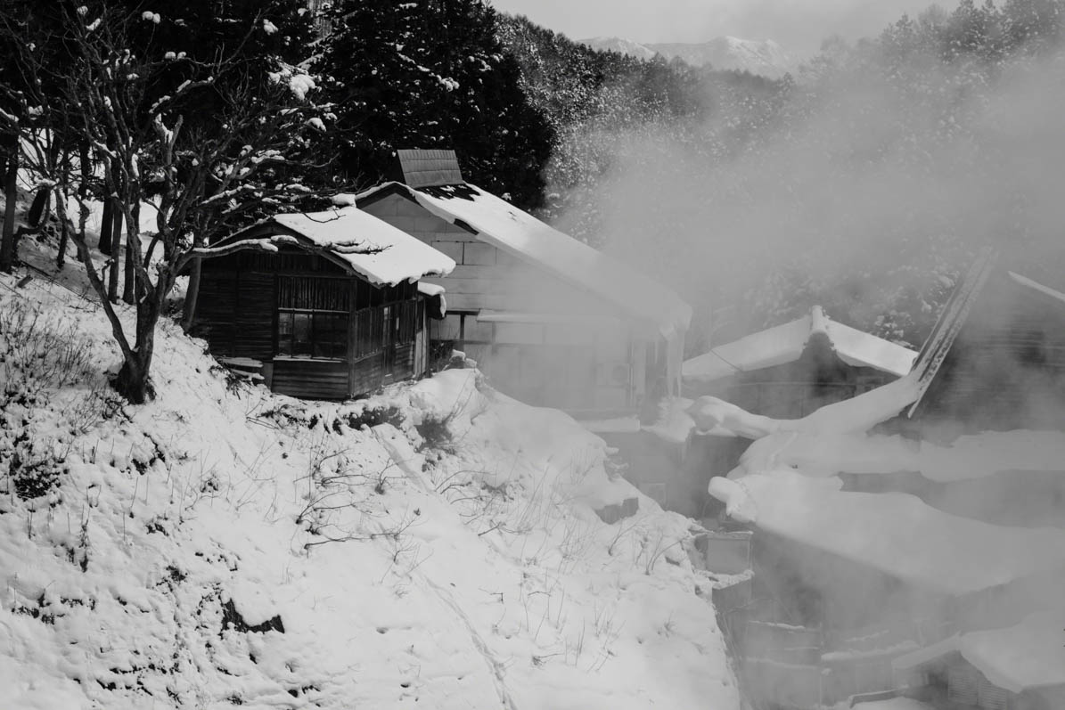 Volcanic spring shooting a plume of hot steam in snow covered hillside, with old mountain huts in Joshinetsu-Kogen National Park, Nagano, Japan